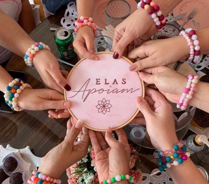 Mãos de mulheres com pulseiras coloridas segurando um bordado da campanha Elas Apoiam